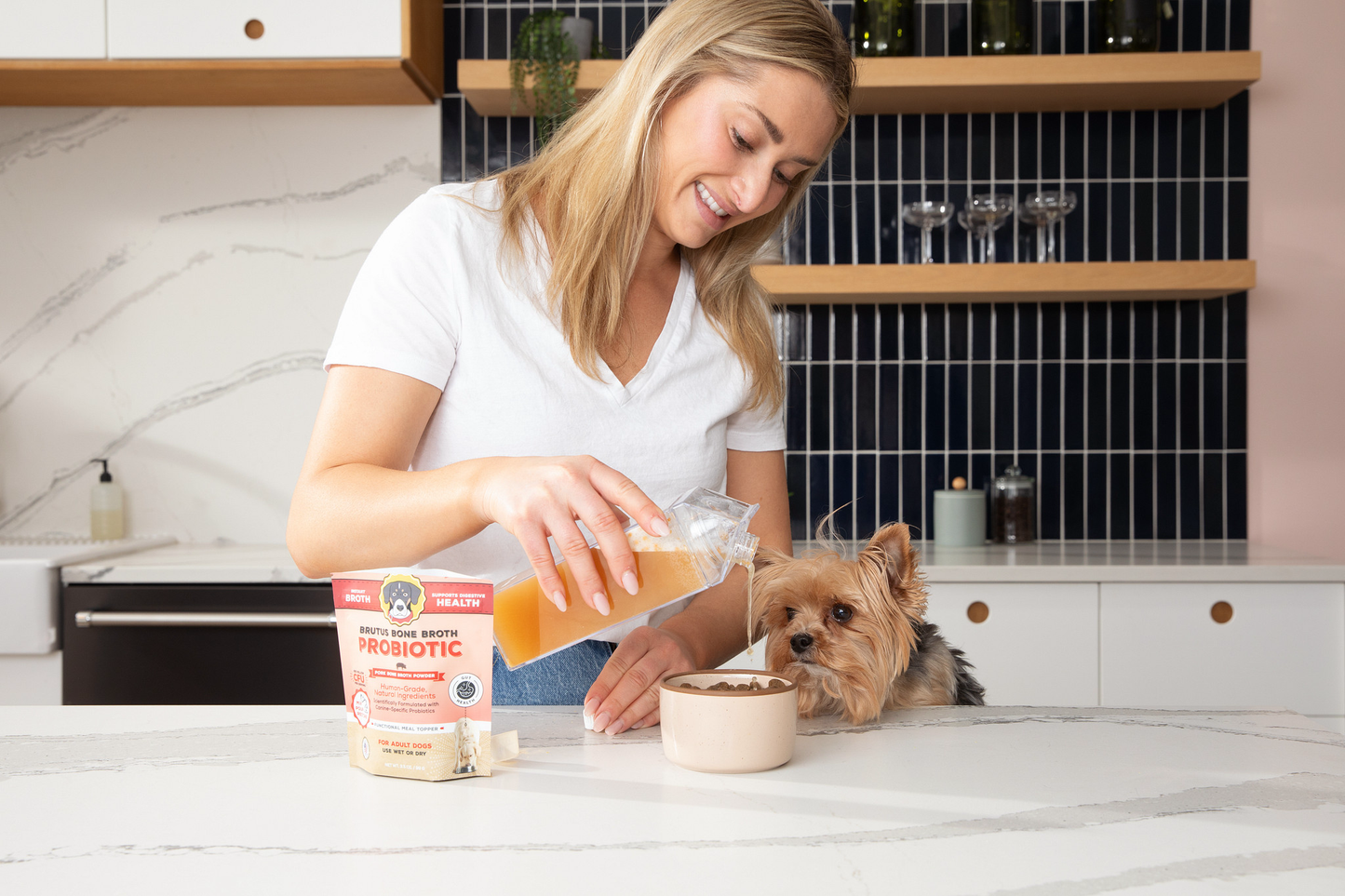 Woman pouring Brutus Broth Probiotic Meal Topper on kibble with a Brown Yorki Dog sitting next to her