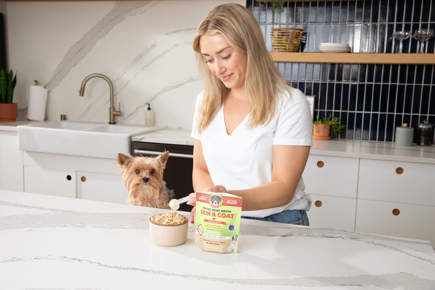 Woman pouring Brutus Broth Skin and Coat Vegetable on kibble with a Brown Yorkie Dog sitting next to her