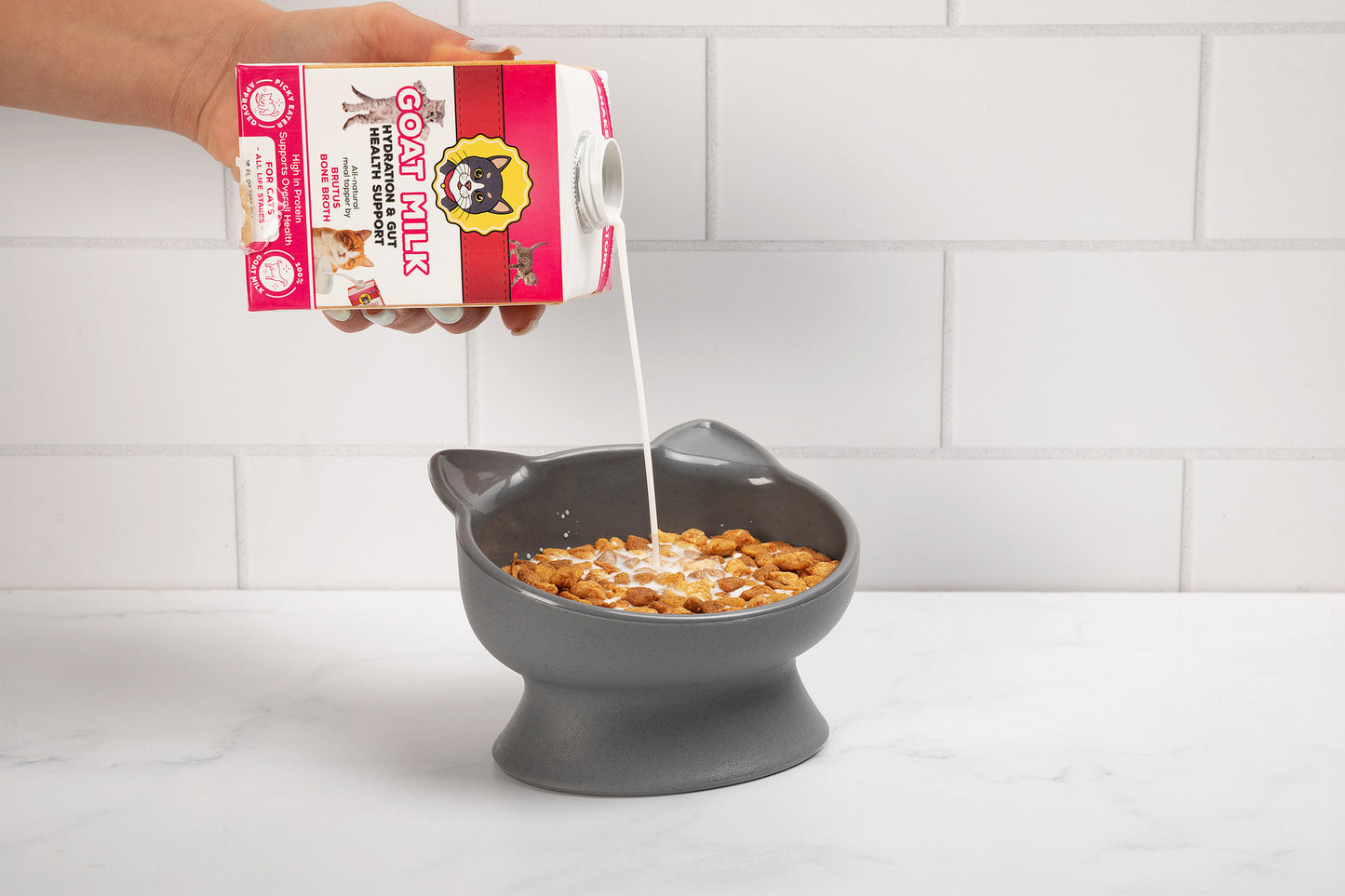 Hand pouring a carton of goat milk labeled "Goat Milk for Cats" into a gray cat-shaped bowl of kibble on a white tiled counter