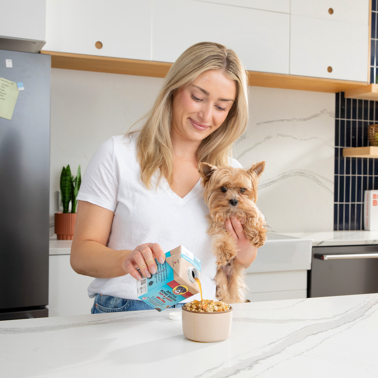 Woman pouring Brutus Broth Hip and Joint Beef on kibble while holding a Brown Yorki Dog