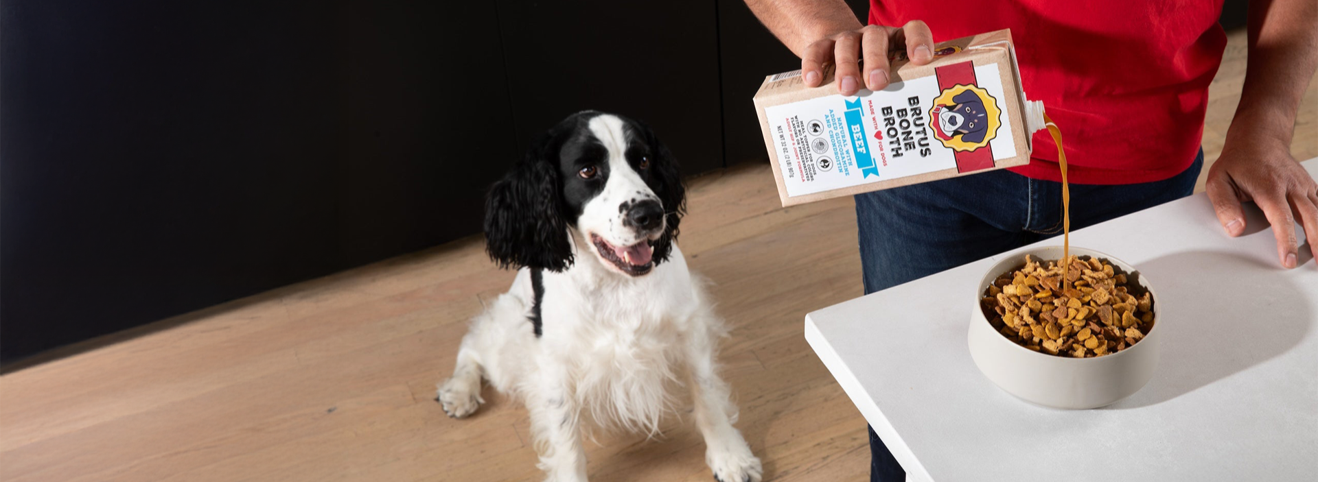 Dog standing next to a person pouring food from a bag into a bowl on a wooden floor.