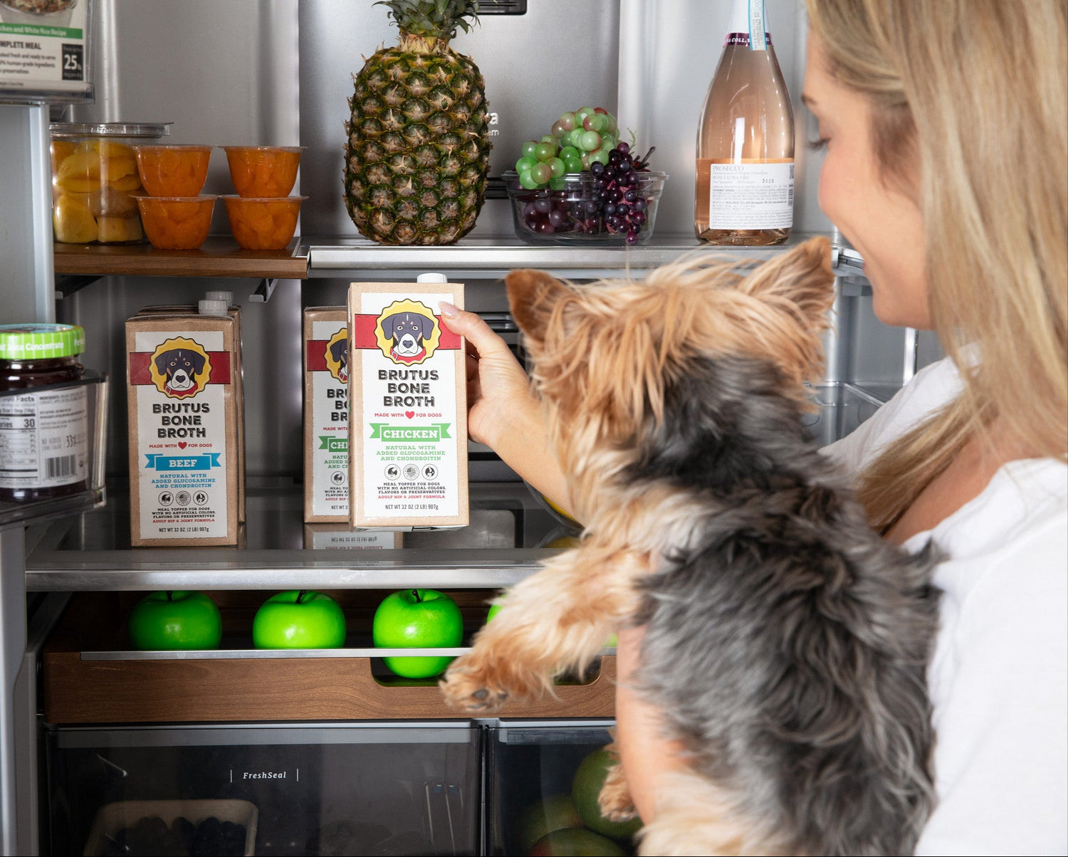 Woman holding a small black and brown dog while pulling a box of Chicken Brutus Bone Broth off the shelf of the fridge.