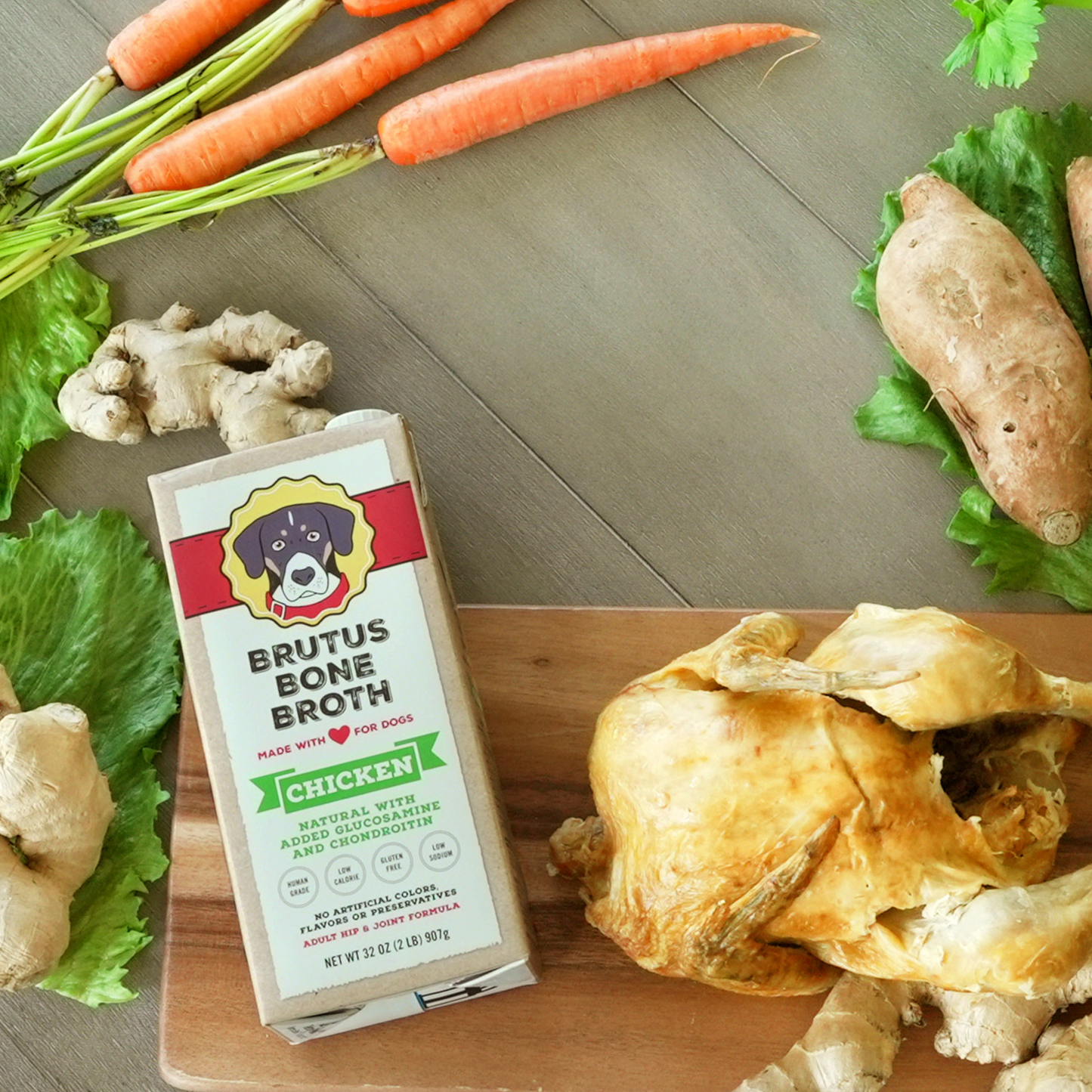 Chicken Brutus Bone Broth box lying on a cutting board with a cooked chicken and raw carrots, ginger, lettuce, and potatoes around it.