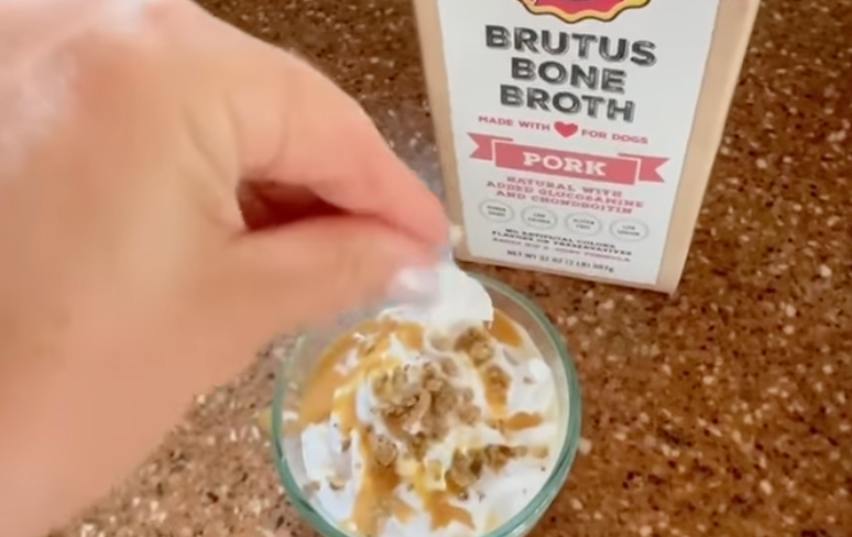 Hand sprinkling crumbled treat onto whipped 'puppaccino' in glass; carton reads: BRUTUS BONE BROTH PORK MADE WITH FOR DOGS