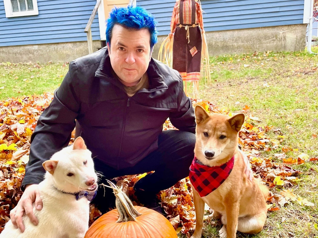 Man crouching in a yard with two dogs amid autumn leaves and a pumpkin, one dog wearing a red plaid bandana and the other a bow tie