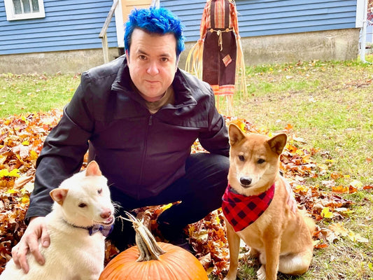 Man crouching in a yard with two dogs amid autumn leaves and a pumpkin, one dog wearing a red plaid bandana and the other a bow tie