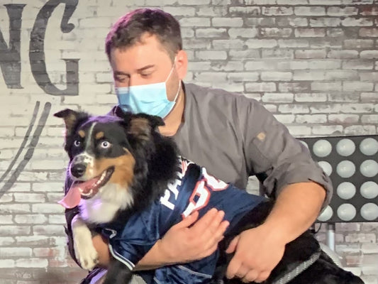 Man wearing a face mask holding an excited black-and-tan dog in a blue football jersey against a brick wall