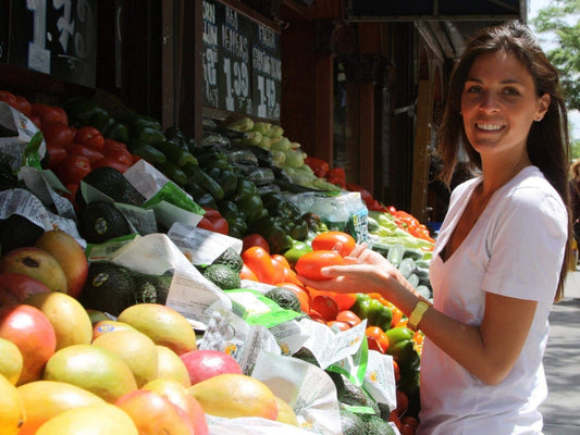 Woman smiling at a farmers' market selecting a tomato from a produce stand with avocados, mangoes and peppers.