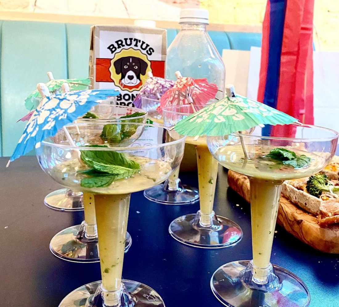 Four cocktail glasses with green creamy drink, mint leaves and paper umbrellas; BRUTUS BONE BROTH carton visible in background
