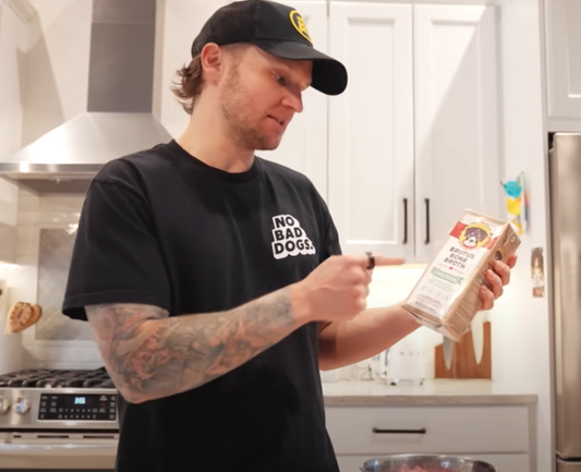 Man in a baseball cap pointing at a carton of Brutus Bone Broth in a bright home kitchen