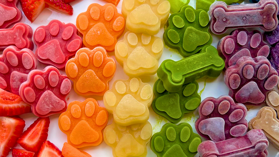 Frozen homemade dog treats shaped like paws and bones in rainbow colors, with sliced strawberries at left
