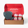 A brown cardboard gift box with an open red interior, containing a dark gray fish-shaped silicone lick mat and two cartons of Goat Milk for Cats with pink labels. The inside lid shows white text reading 'GIVE THEM THEIR BEST DAY, EVERY DAY.'