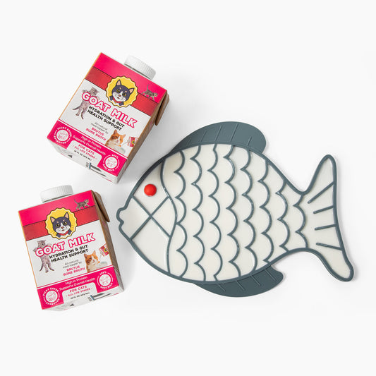 A dark gray fish-shaped silicone lick mat filled with a Goat Milk, shown next to two cartons of Goat Milk for Cats on a clean white background.