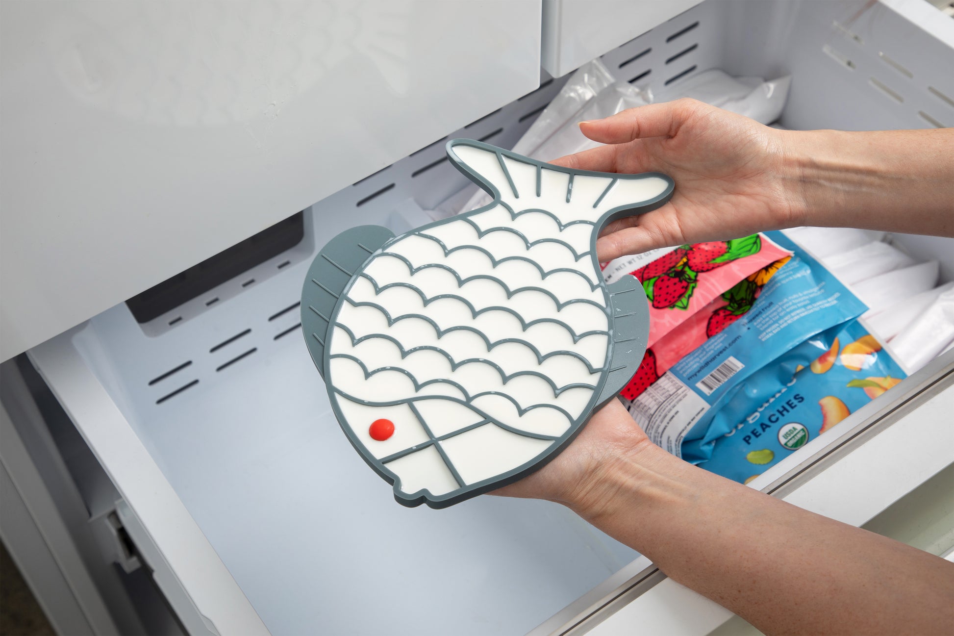 A person's hands holding a grey fish-shaped silicone lick mat filled with Goat Milk and a small red accent, positioned in front of an open freezer drawer containing packaged frozen foods.