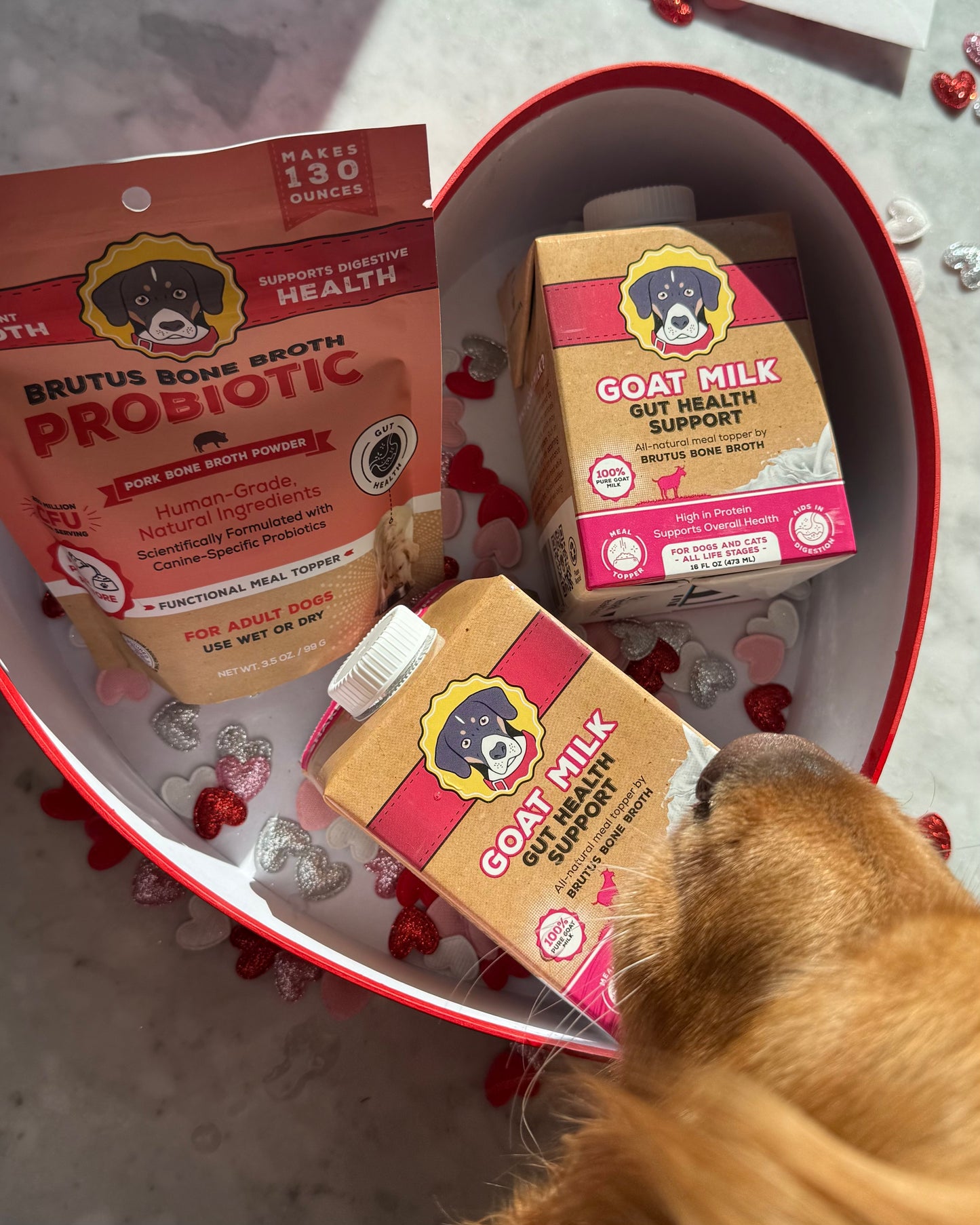 Brutus Bone Broth products including probiotic powder and goat milk supplement in a heart-shaped container with a dog's paw reaching in.